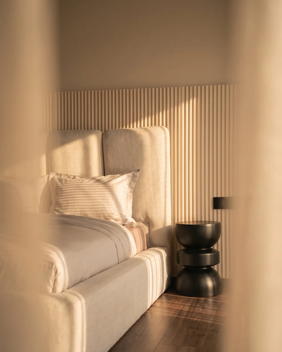 La suite parentale de la villa témoin Jnane Oum El Ghite au couchant — tête de lit capitonnée crème, mur cannelé baigné de lumière dorée, chevet noir et parquet en noyer, photographiée sur place.