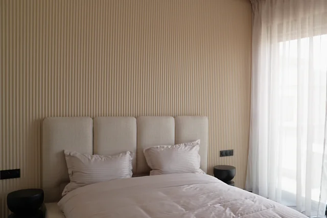La suite parentale de la villa témoin : mur cannelé beige, tête de lit capitonnée, voilages devant la baie — vue d’ambiance.