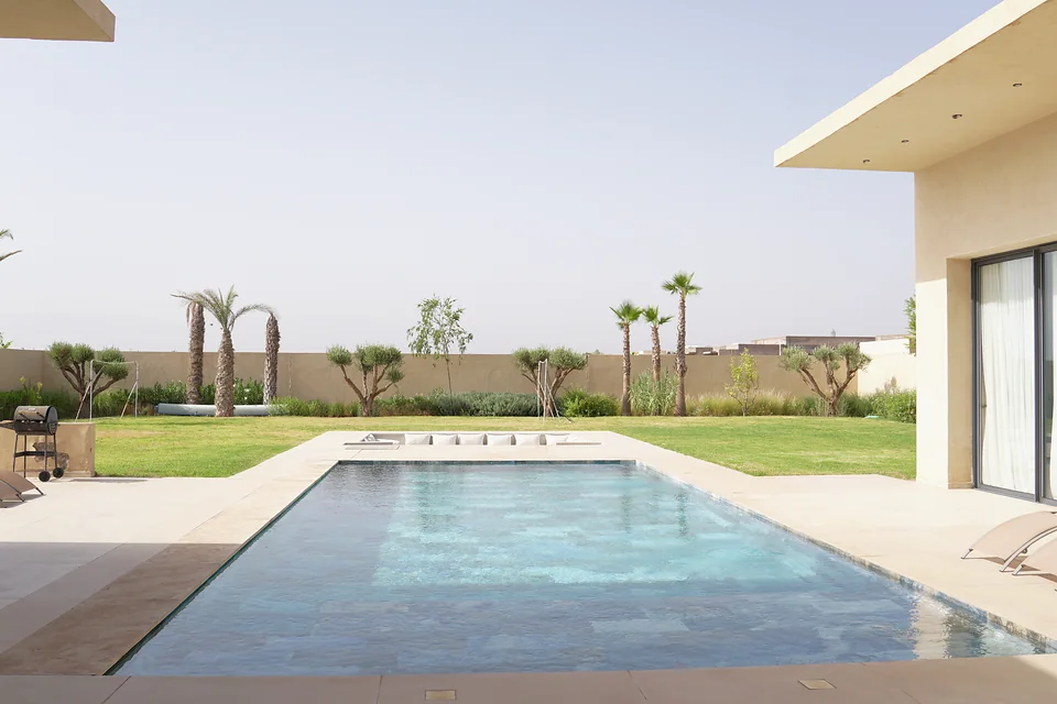 Le jardin de la villa témoin Jnane Oum El Ghite : le bassin dans sa longueur, la pelouse jusqu’au mur d’enceinte, palmiers et oliviers, et la villa et ses baies toute hauteur à droite — photographié sur place.